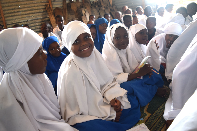 Young girls wearing white hijabs and blue skirts (their school uniforms) are smiling at the camera