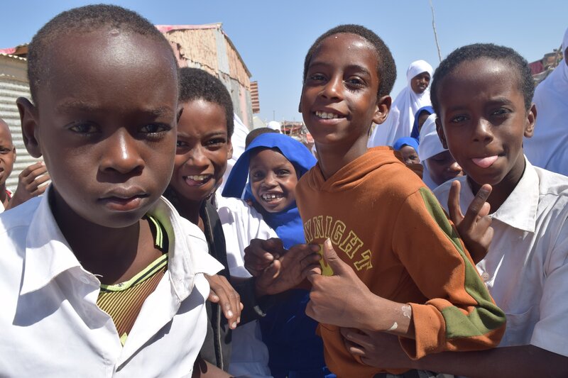 Young boys are smiling at the camera, there are also a few young girls in the background looking into the camera from behind them