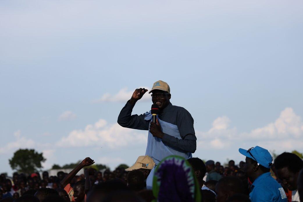 Person holding a microphone and papers while addressing a large outdoor crowd under a clear sky.